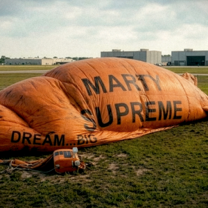 Deflated orange blimp branded “Marty Supreme” lying on a grassy airfield with two people standing nearby, suggesting a failed or grounded marketing campaign.