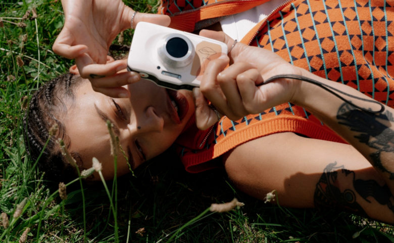 Person lying in tall grass, taking a photo with a white compact camera, wearing an orange patterned garment and showing tattoos on the forearm.