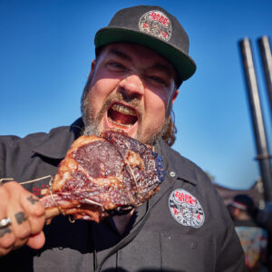 Man wearing a dark cap with a logo bites into a large piece of grilled meat outdoors against a blue sky.