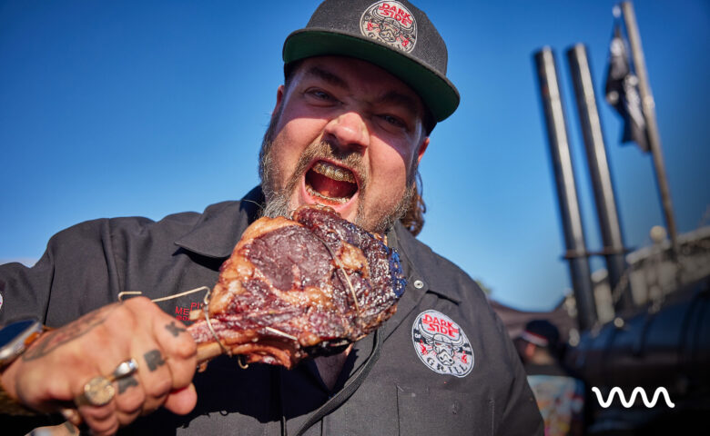Man wearing a dark cap with a logo bites into a large piece of grilled meat outdoors against a blue sky.