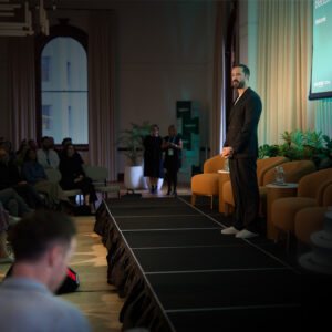 Bearded man in a dark suit stands on stage delivering a talk to an audience in a dim conference room.