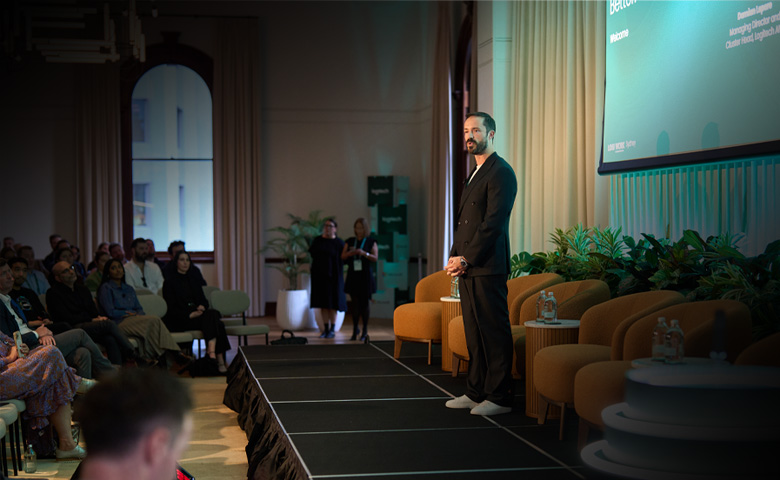 Male presenter in a black suit on a stage speaking to an audience in a conference room with a large screen to the right and green plants nearby.